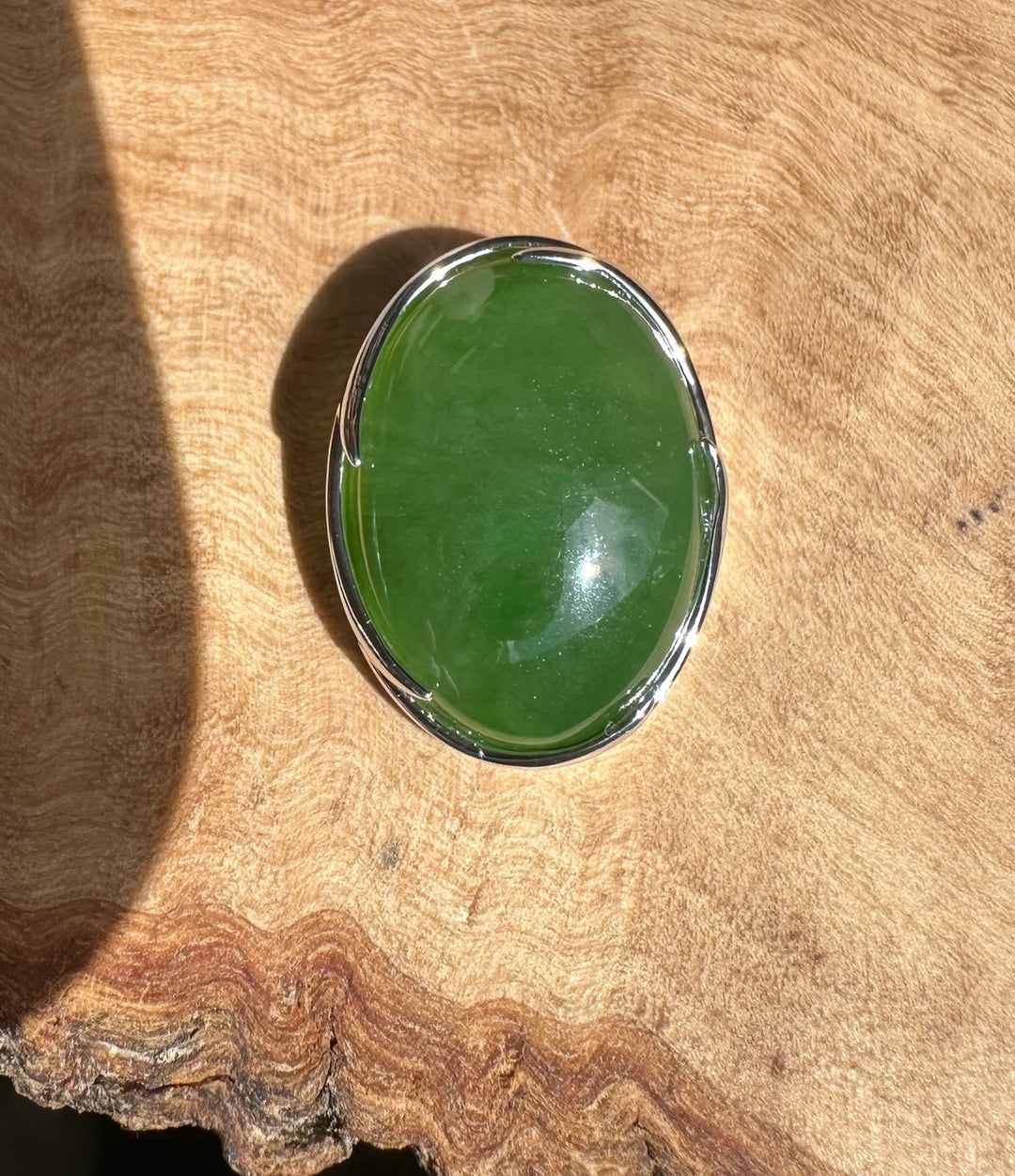 Oval bright green Canadian jade pendant in a silver frame resting on textured wood.