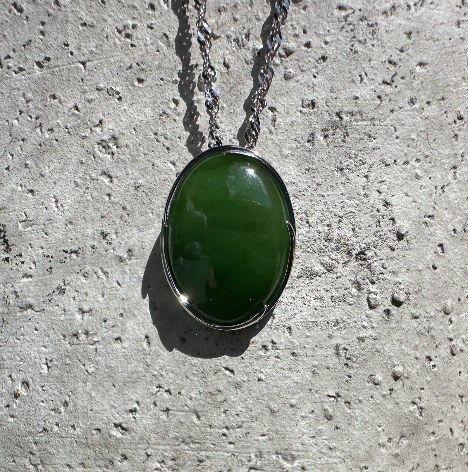 Oval bright green jade pendant in a silver bezel with chain against a concrete backdrop
