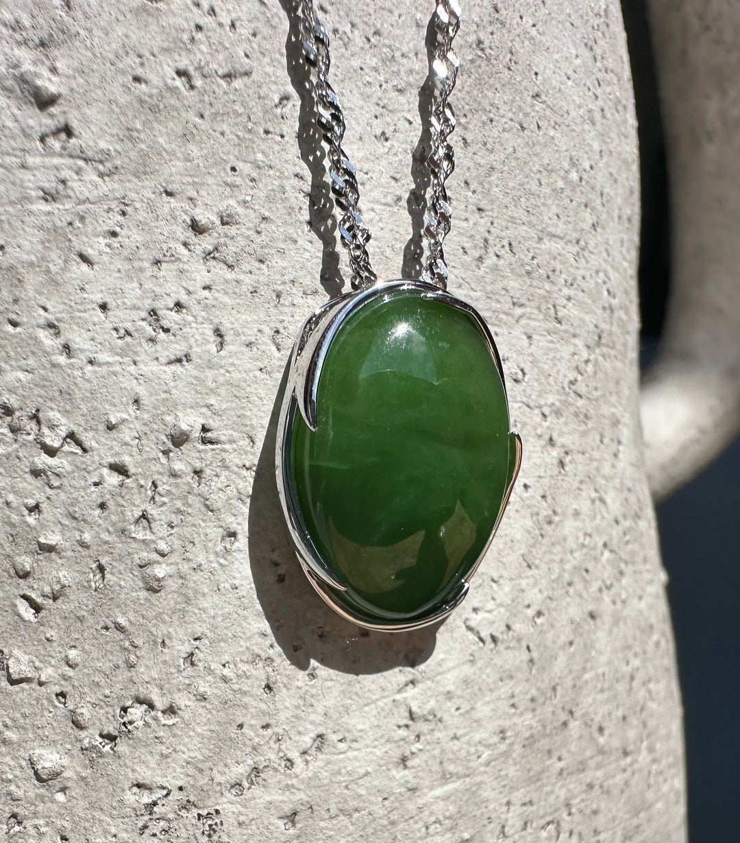Bright green Canadian jade pendant in a silver bezel on a twisted silver chain against a light stone background.