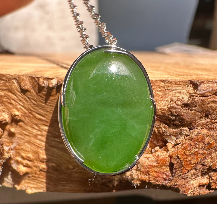 Close up of an oval bright green jade pendant in a silver setting on sunlit wood