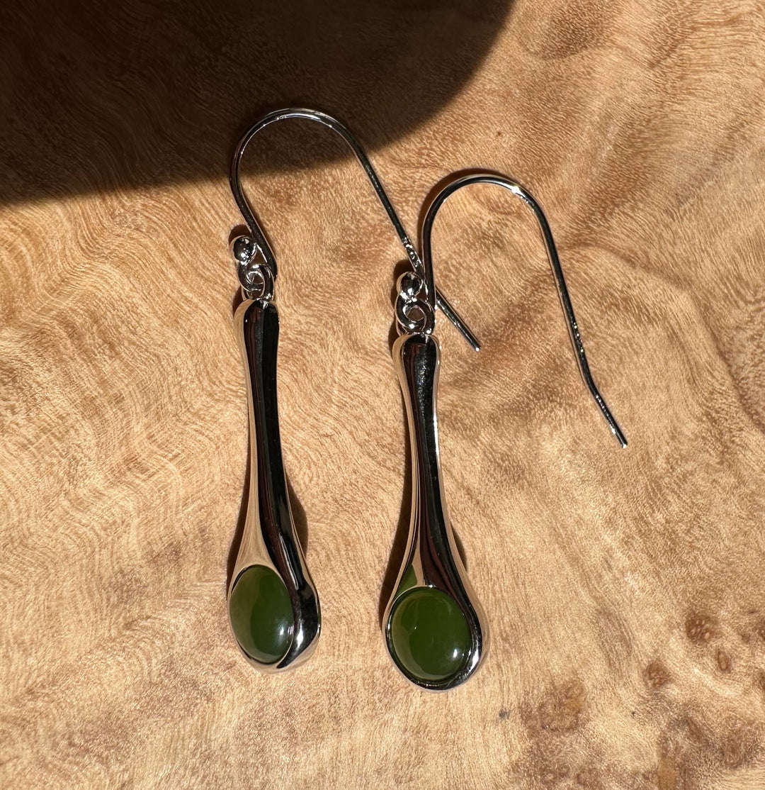 Silver drop earrings with elongated curved bodies and green round stones on a wooden surface