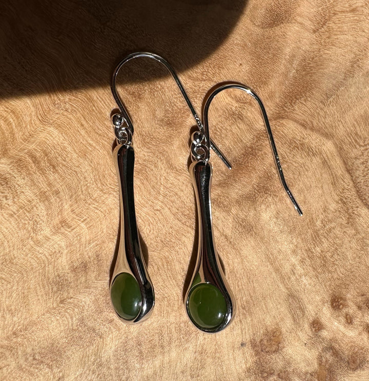 Silver drop earrings with elongated curved bodies and green round stones on a wooden surface