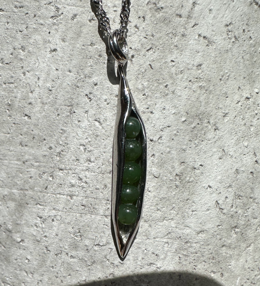 Close up of a jade peapod pendant in stainless steel with green beads visible inside against a textured stone background