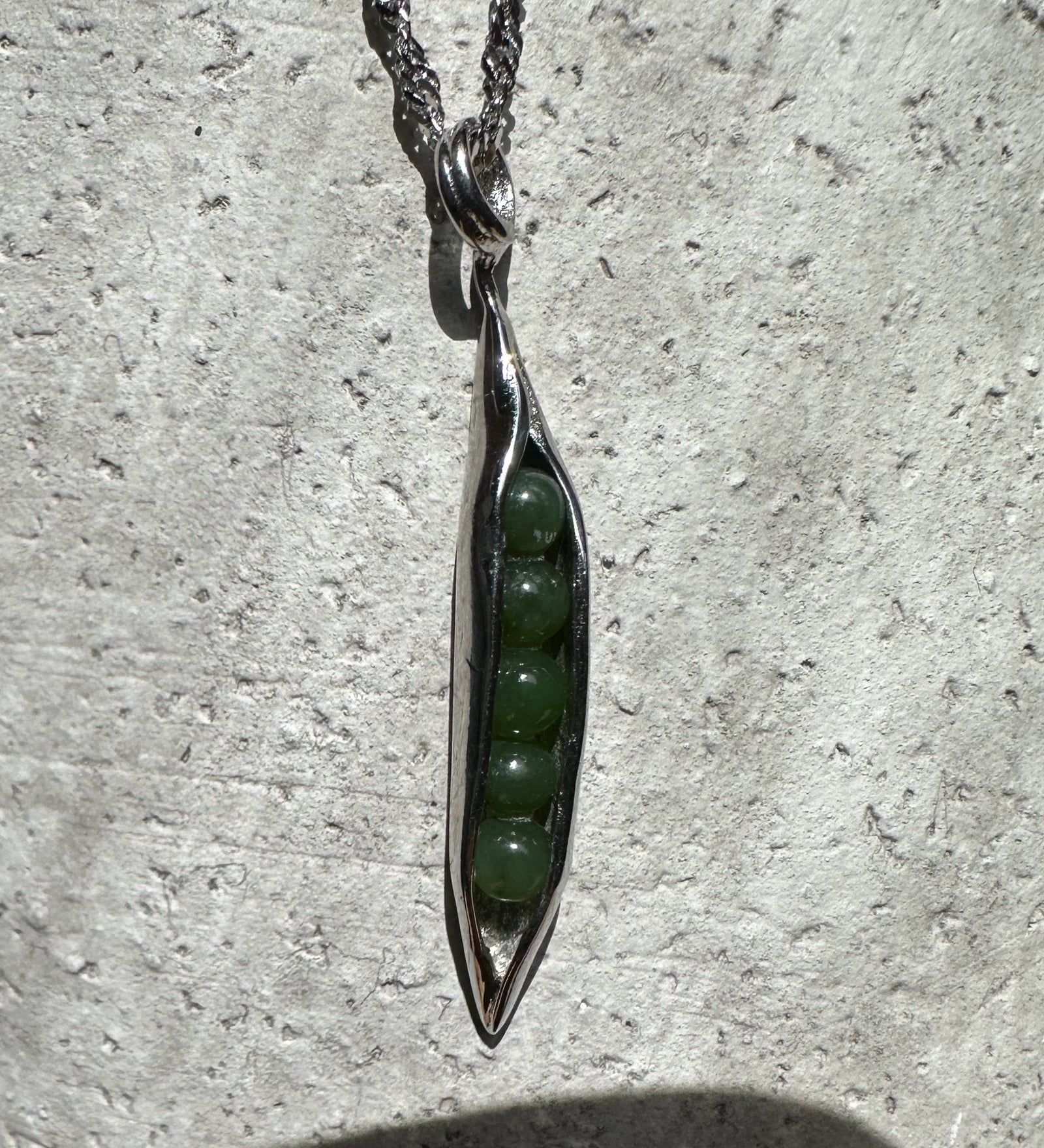Close up of a jade peapod pendant in stainless steel with green beads visible inside against a textured stone background
