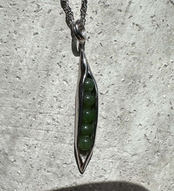 Close up of a jade peapod pendant in stainless steel with green beads visible inside against a textured stone background