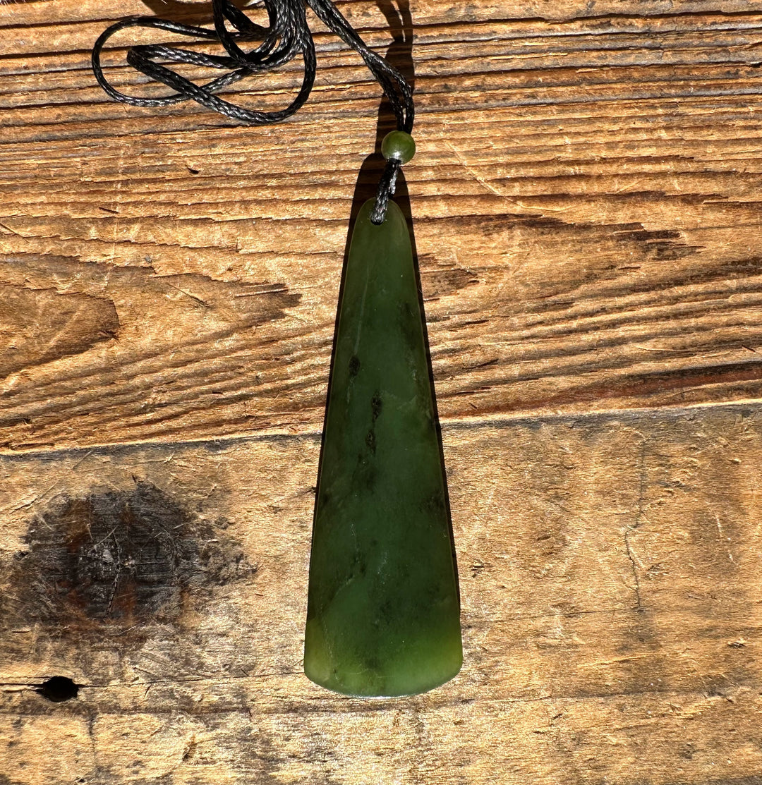 Green jade drop pendant on a black cord with a small bead, shown on a wooden surface.
