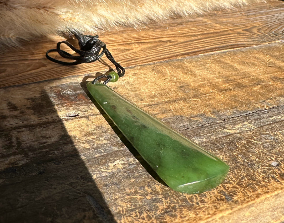 Green jade drop pendant on a knotted black cord with a small bead, lying on a rustic wooden surface