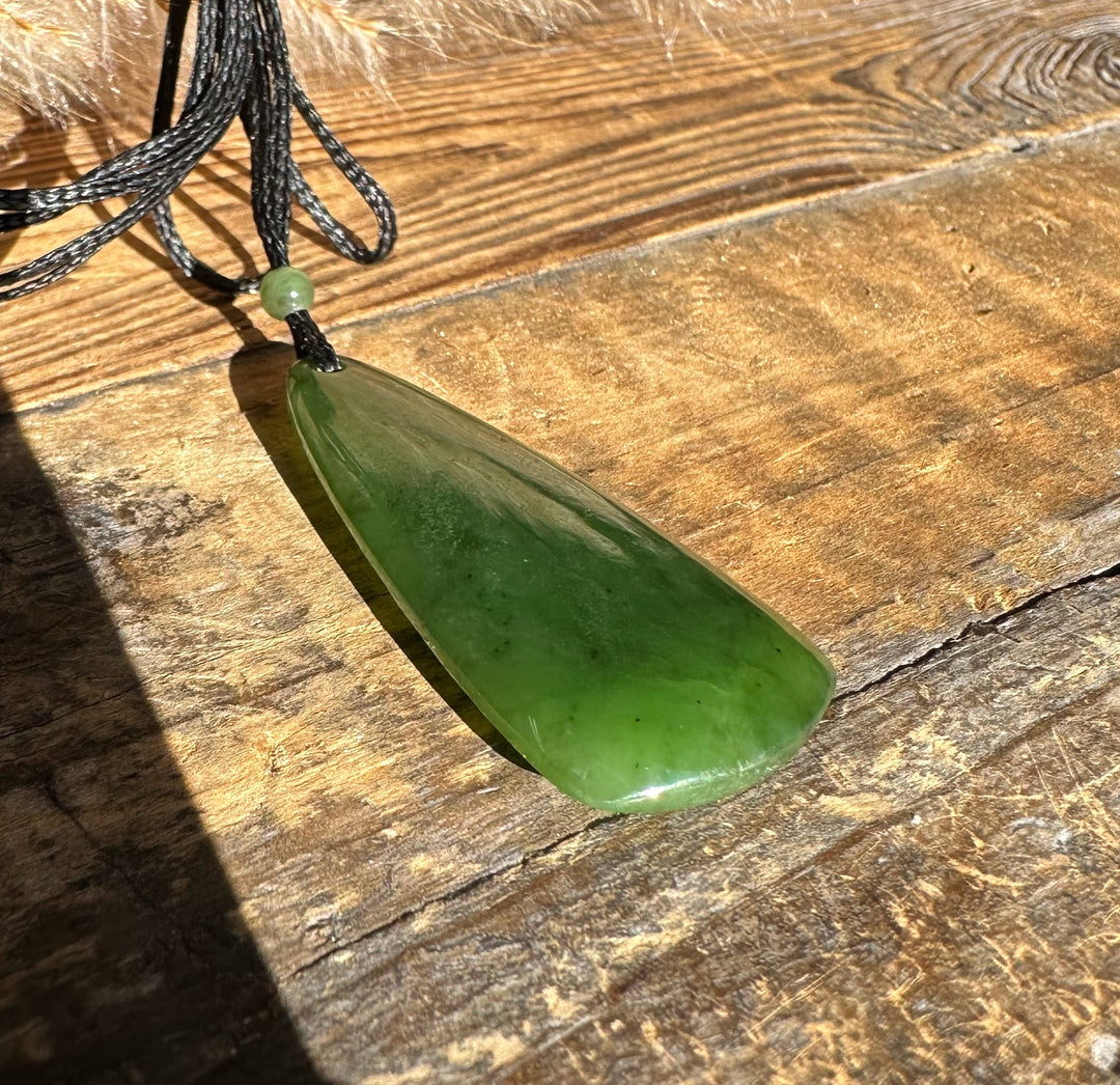 Green jade teardrop pendant on black cord with a small bead above, laid on a wood surface