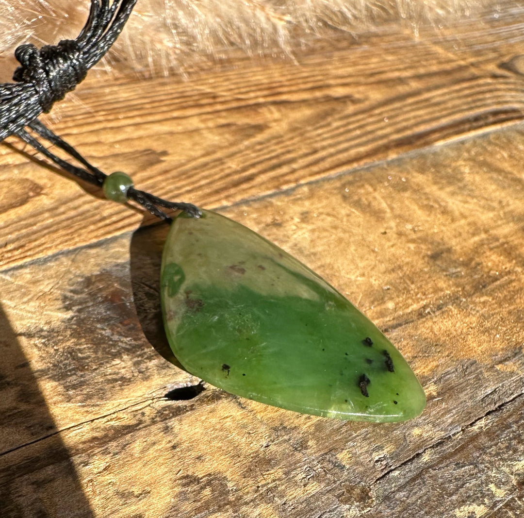 Green jade pendant shaped like a rounded leaf on a black cord, displayed on a rustic wooden surface.