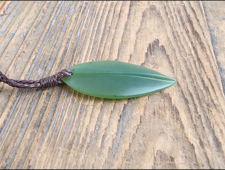 Green jade leaf pendant resting on a wooden surface with its brown cord nearby.