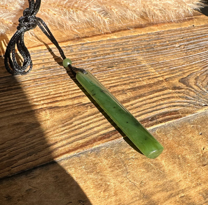 Green jade cylinder pendant on a black cord with a bead, laid on a wooden surface
