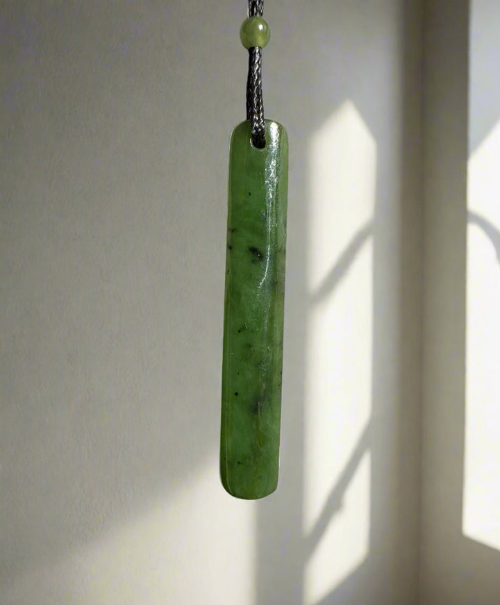 Close-up of a green jade drop pendant on a black cord hanging against a light colored wall.
