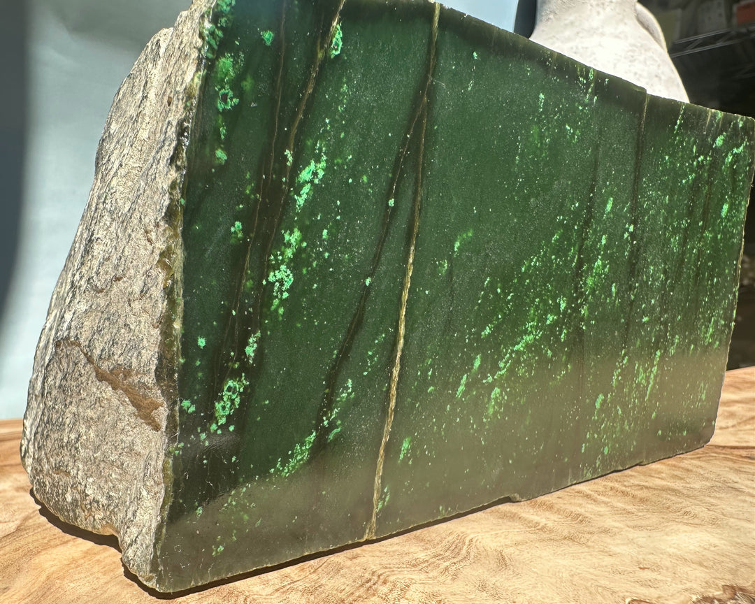 Rough Cassiar jade slab with deep green hue and vivid emerald flecks on a wooden surface