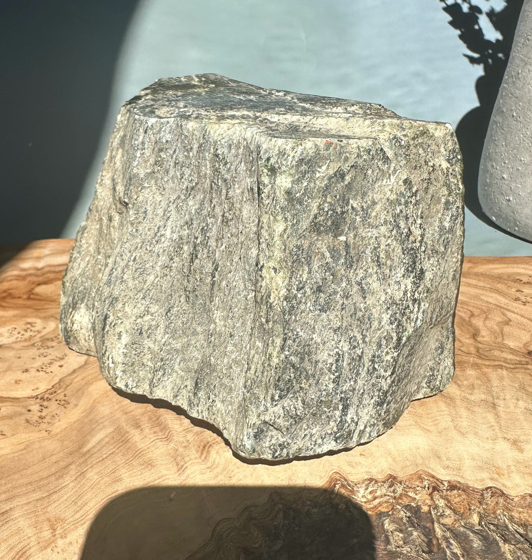 Dark green Cassiar Jade rough rock with vertical striations and light beige patches on a wooden surface.