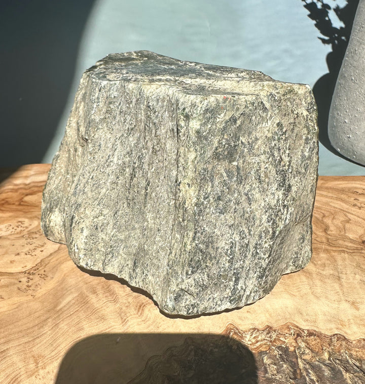Dark green Cassiar Jade rough rock with vertical striations and light beige patches on a wooden surface.