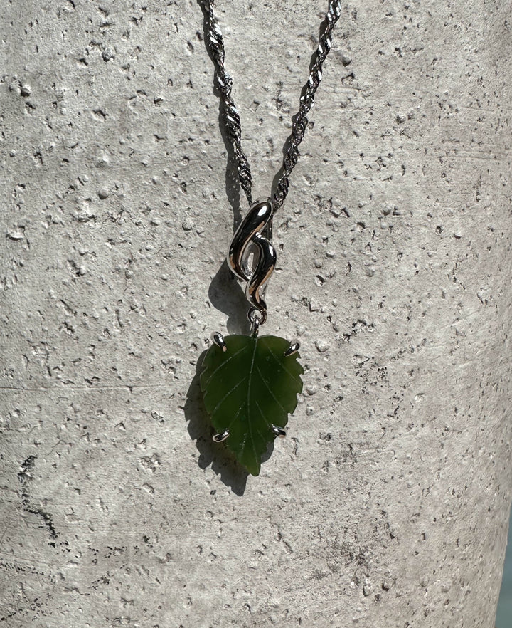 Silver leaf pendant with a green leaf-shaped stone and twisted bail on a textured background.
