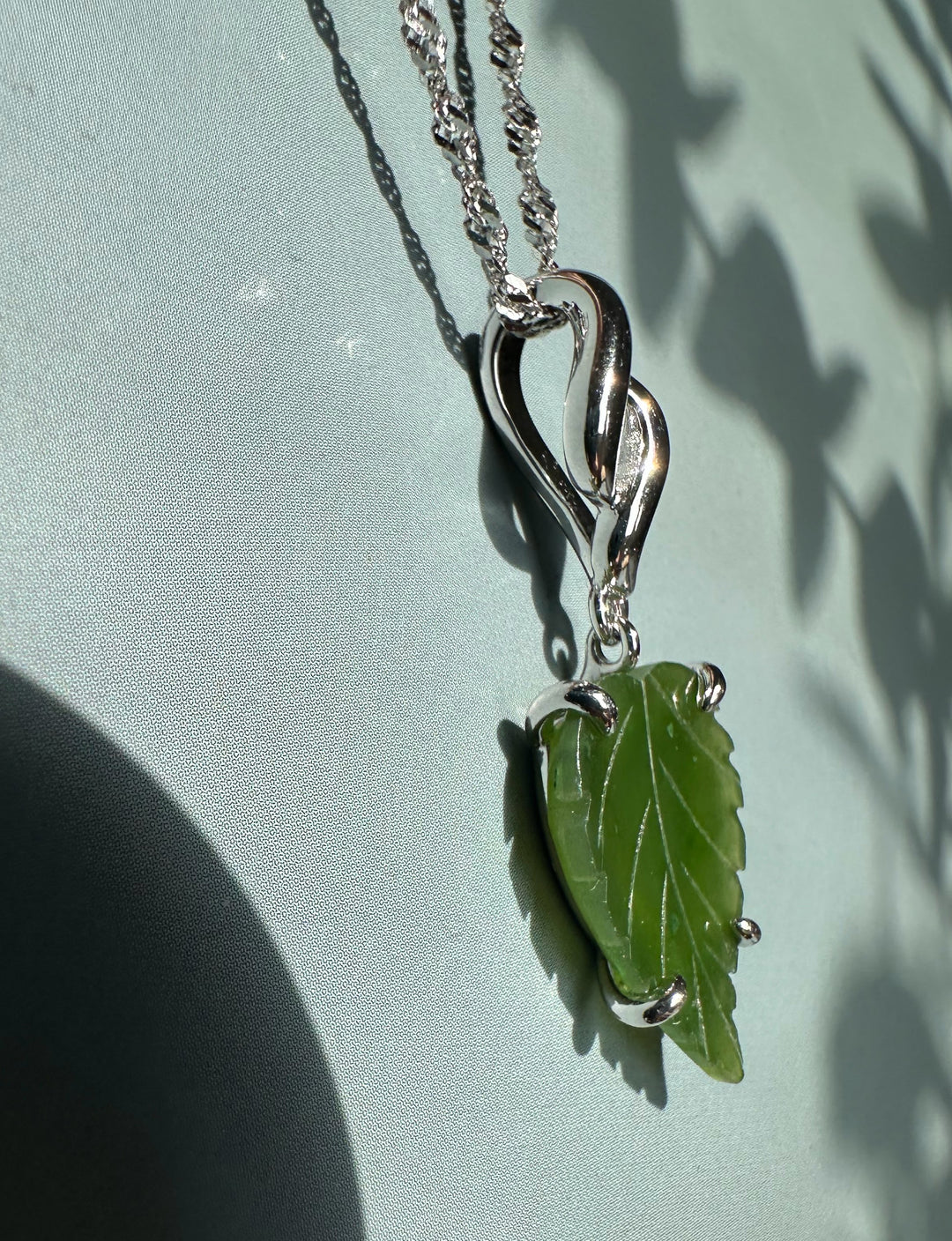 Green leaf pendant with curved silver bail and chain against a pale blue backdrop