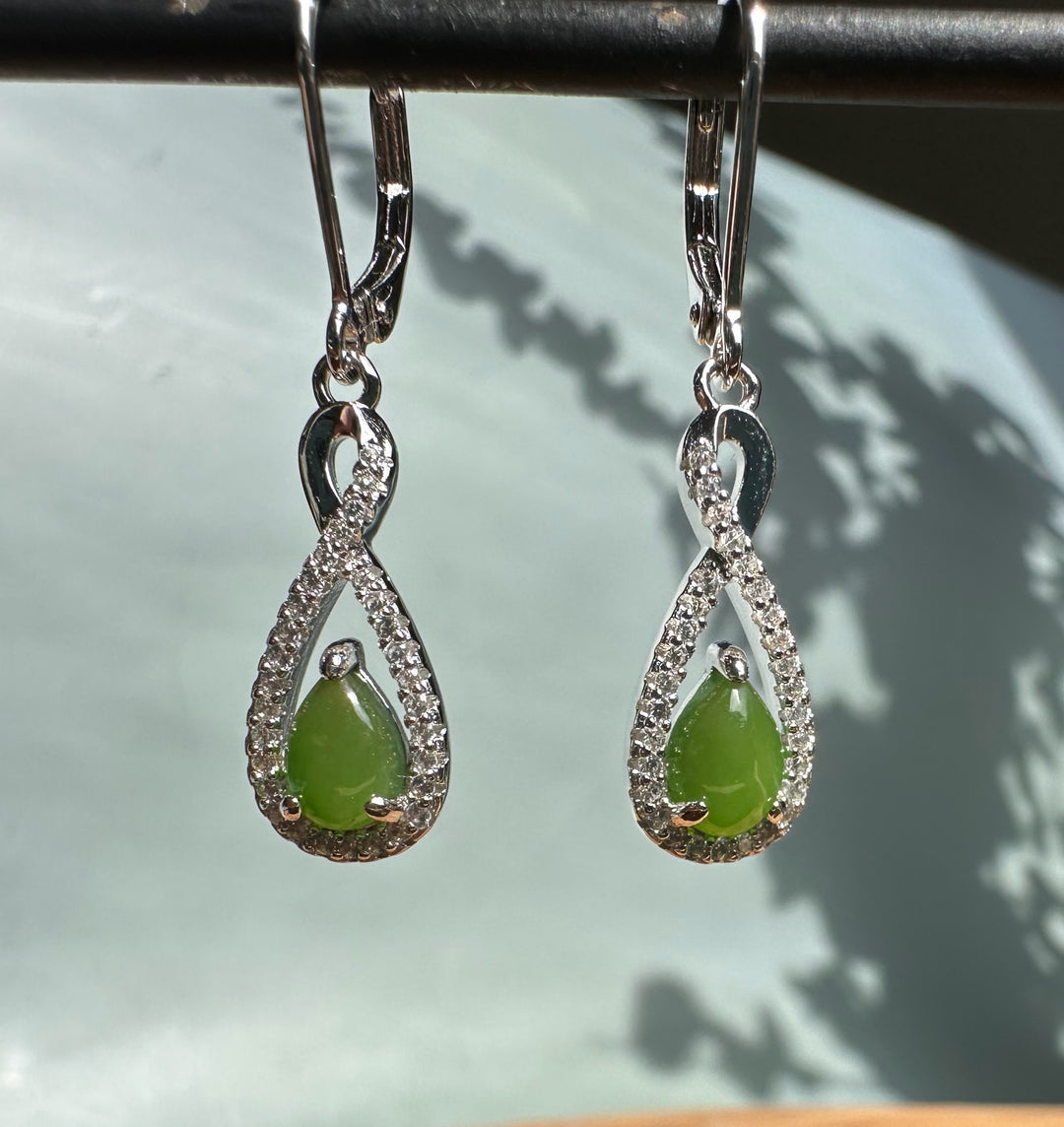 Silver infinity twist earrings with green jade teardrops hanging against a pale backdrop.