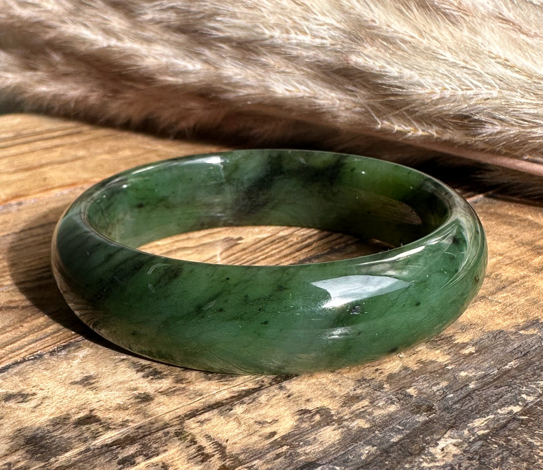 Wide jade bangle 15mm thick in green with marbling and dark specks on a wooden surface