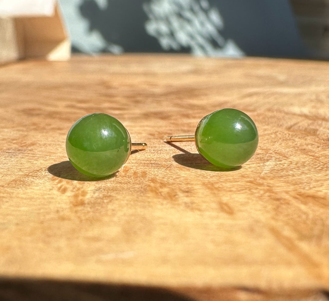 Pair of round green jade stud earrings with gold posts on a wooden surface.
