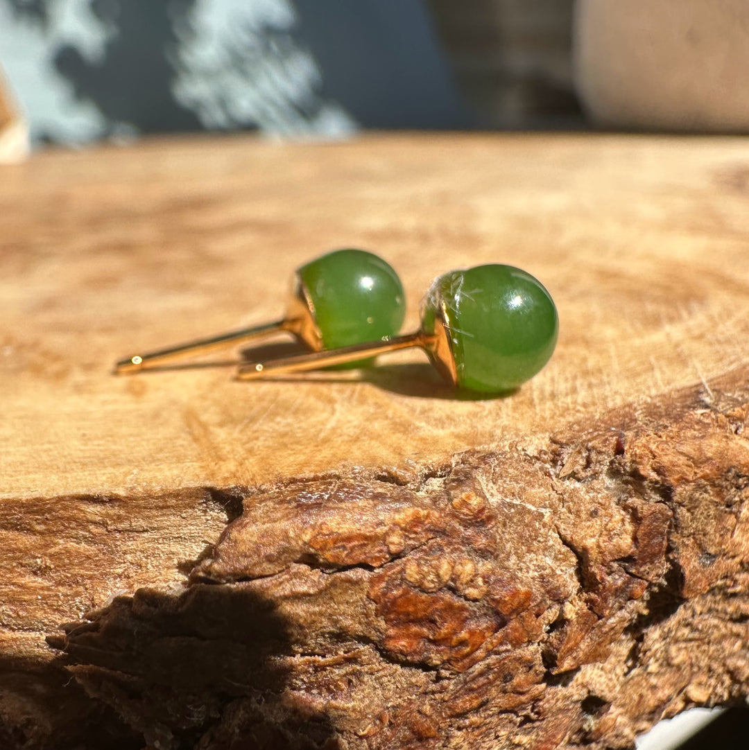 Two round green jade stud earrings with gold posts resting on a textured wooden surface.