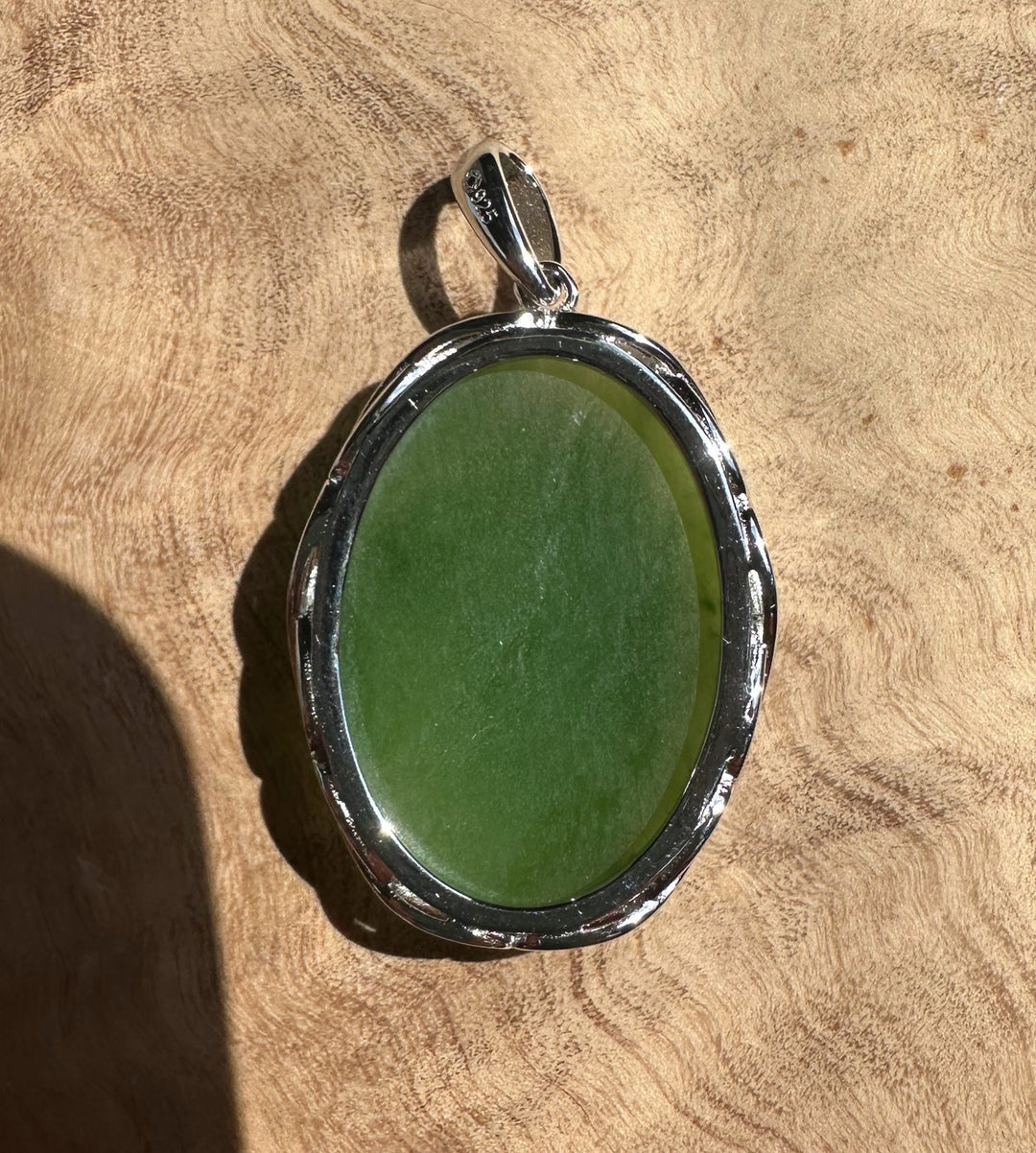 Front view of an oval green jade pendant in a silver setting with a bail against a textured surface