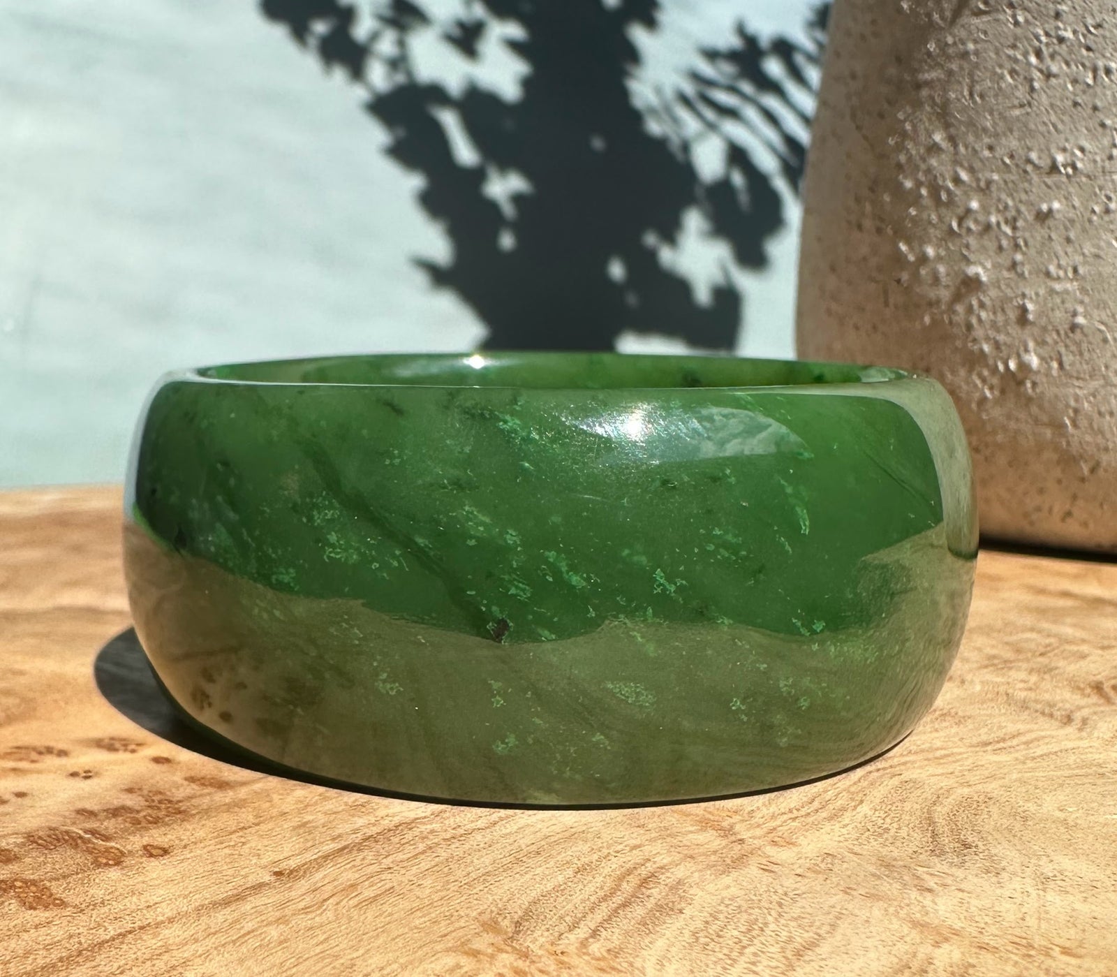 Wide Cassiar jade bangle in deep green with lighter swirls on a wooden surface.