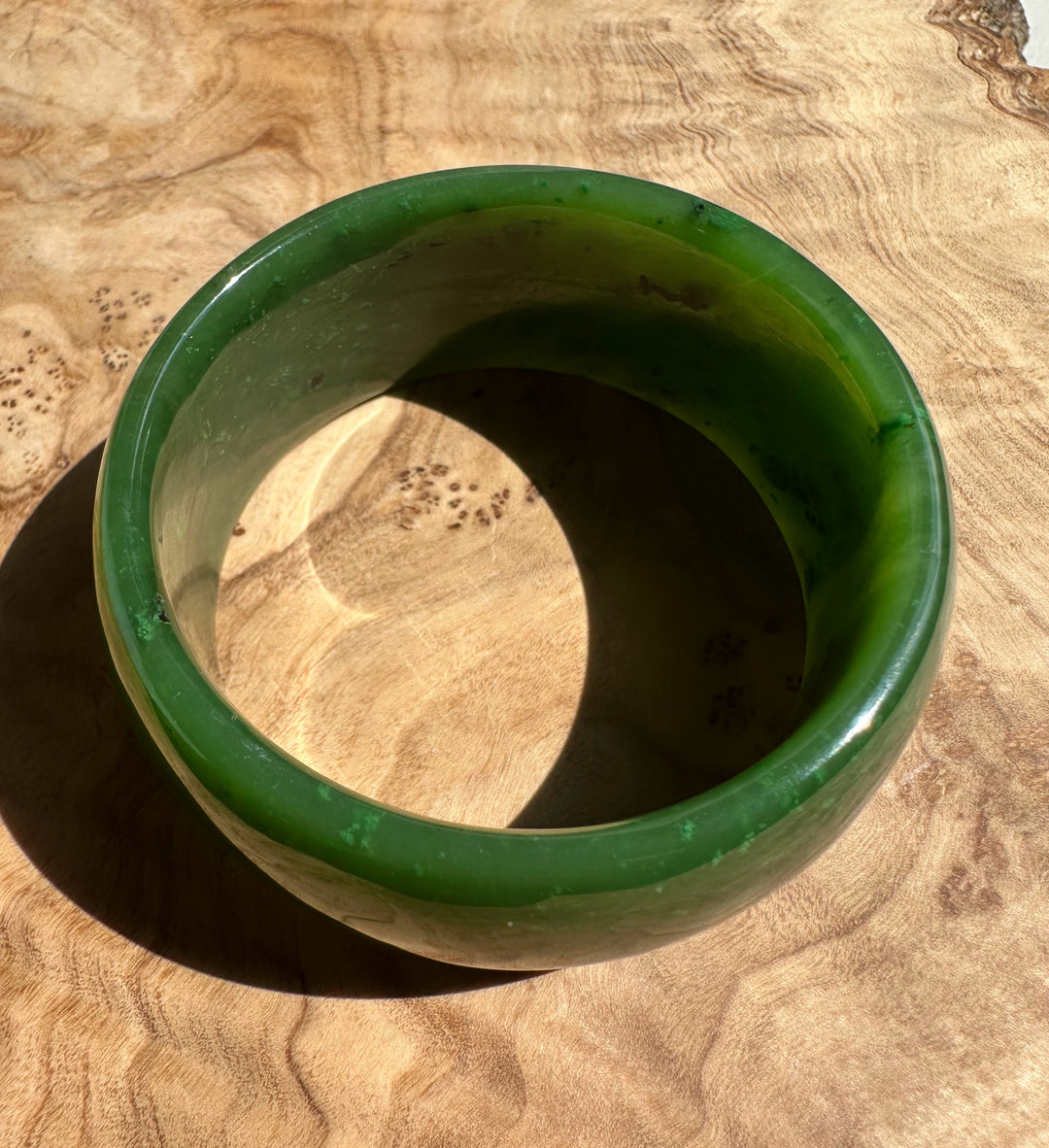 Wide Cassiar Jade Bangle in vivid green resting on a wooden surface.