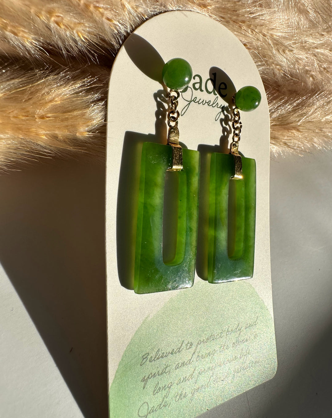 Green jade circle stud connected by a gold chain to a rectangular jade drop on a white display card