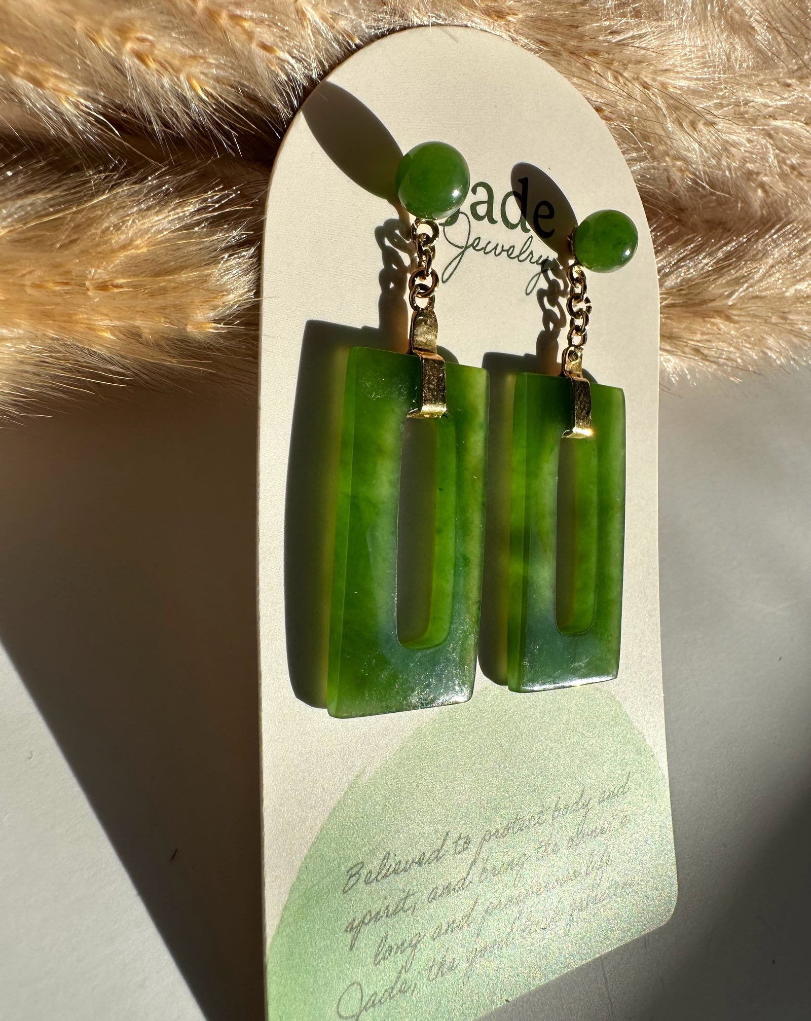 Green jade circle stud connected by a gold chain to a rectangular jade drop on a white display card