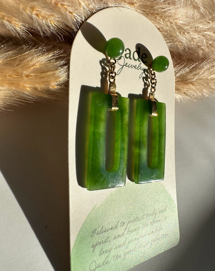 Green jade circle stud connected by a gold chain to a rectangular jade drop on a white display card