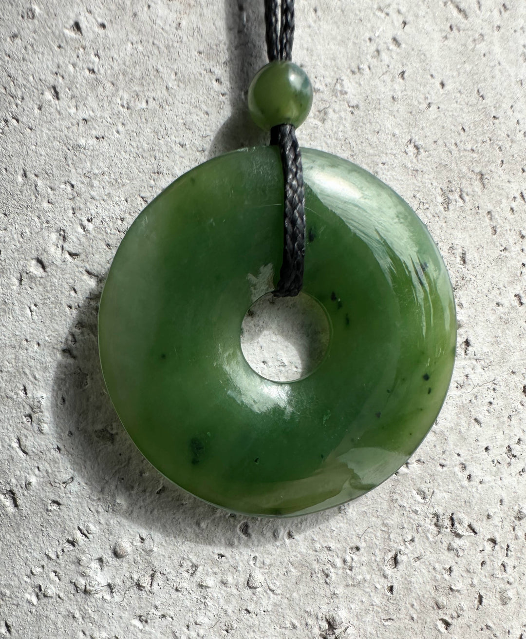 Green jade donut pendant with central hole on black cord and small bead above.