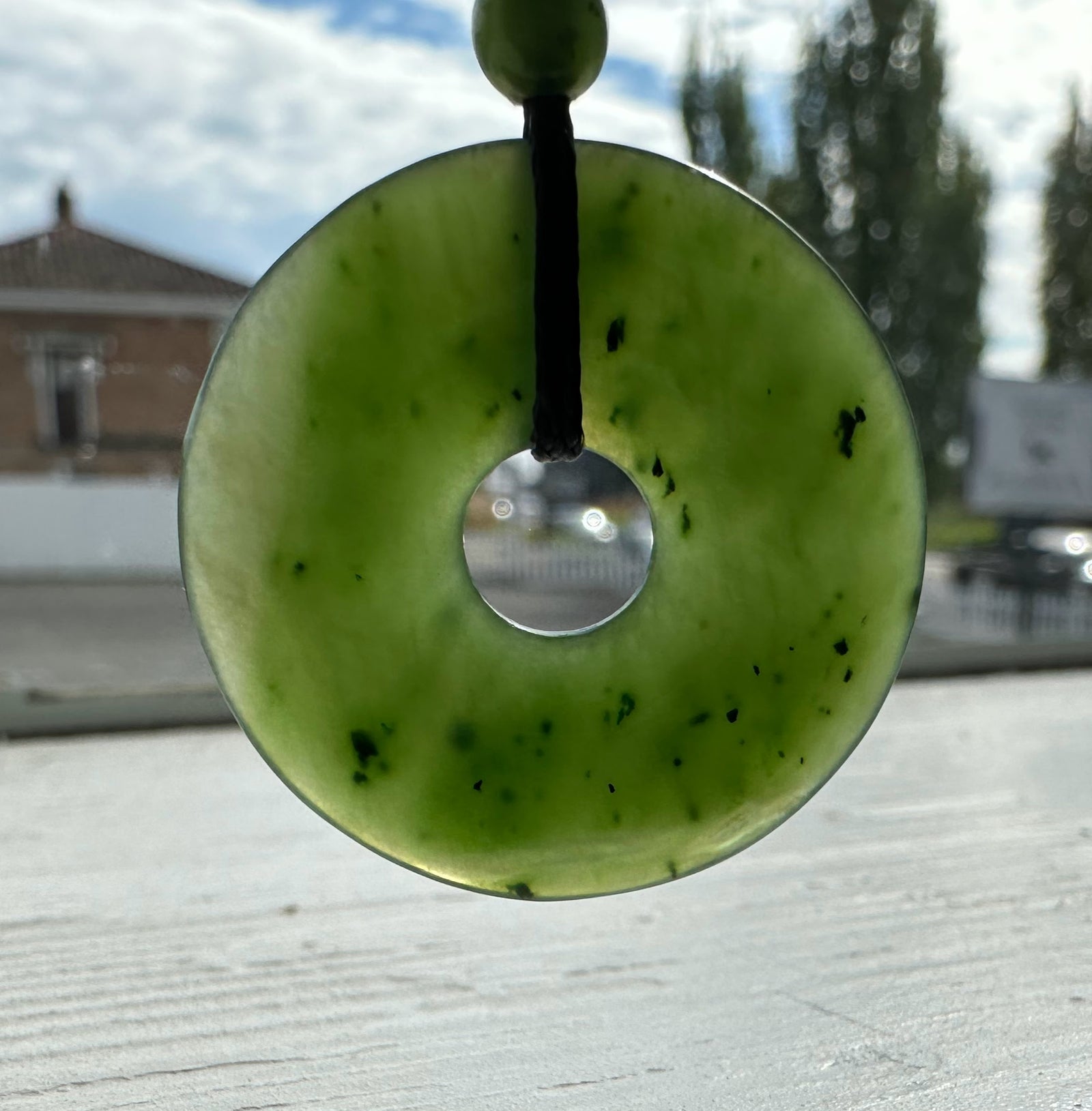 Green jade donut pendant on black cord with bead, photographed outdoors against a blurred urban background.