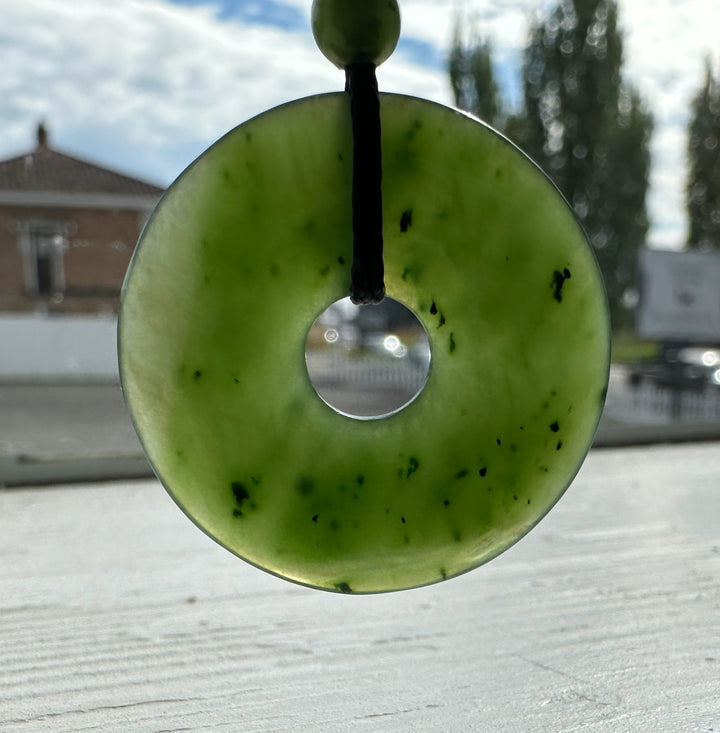 Green jade donut pendant on black cord with bead, photographed outdoors against a blurred urban background.