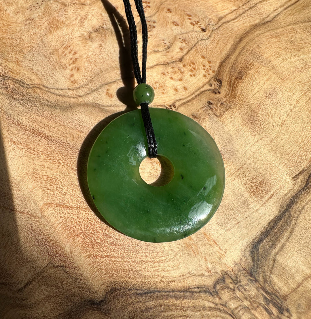 Green jade donut pendant with central hole on black cord and bead, laid on a wooden surface.