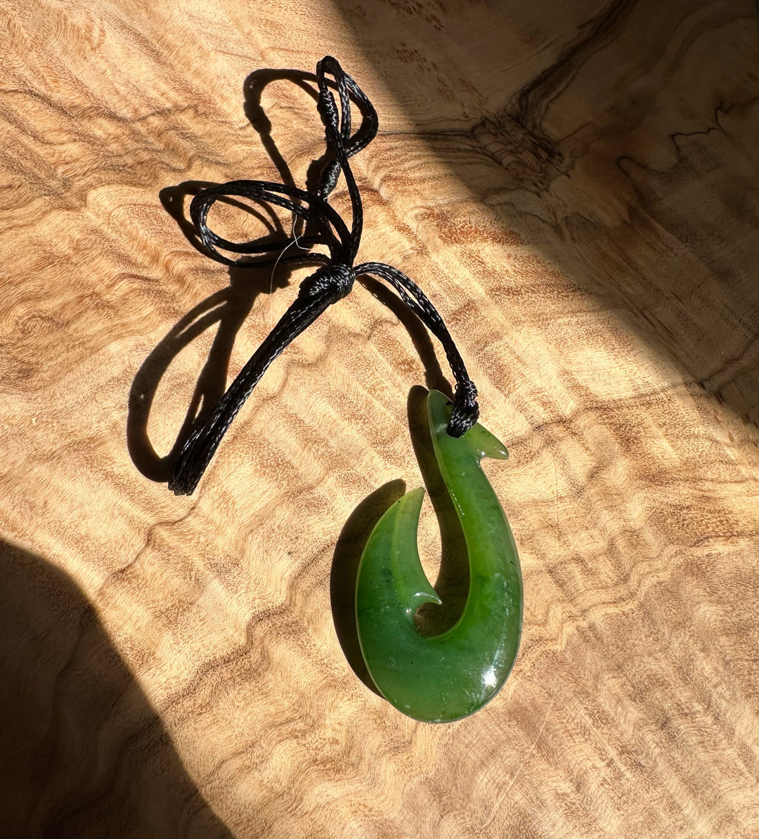 Polished green fish hook pendant tied to a black cord resting on a wooden surface.
