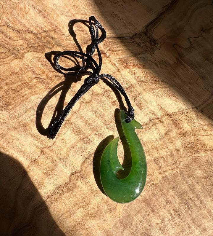 Polished green fish hook pendant tied to a black cord resting on a wooden surface.