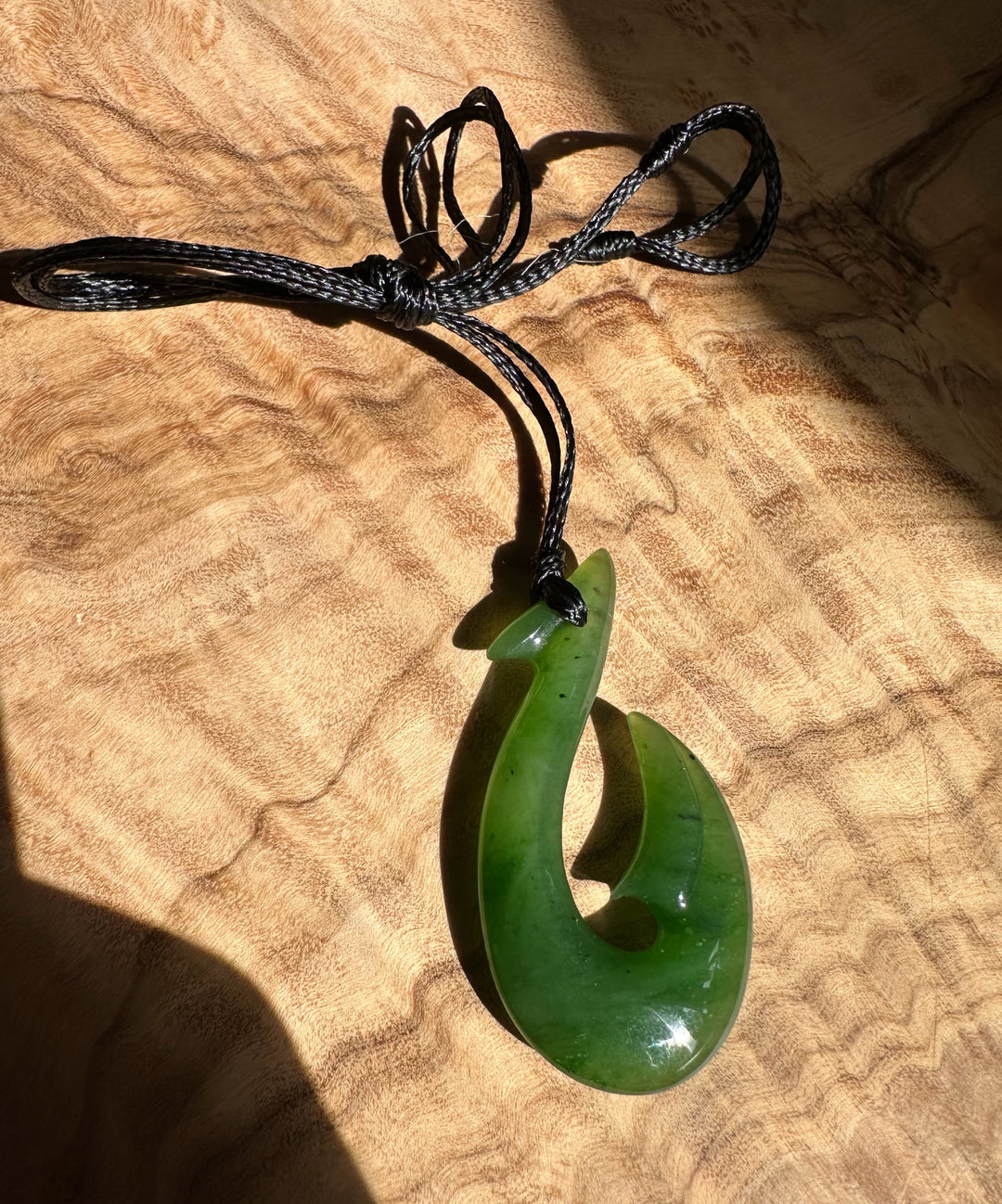 Green jade fish hook pendant on a black braided cord against brown fabric