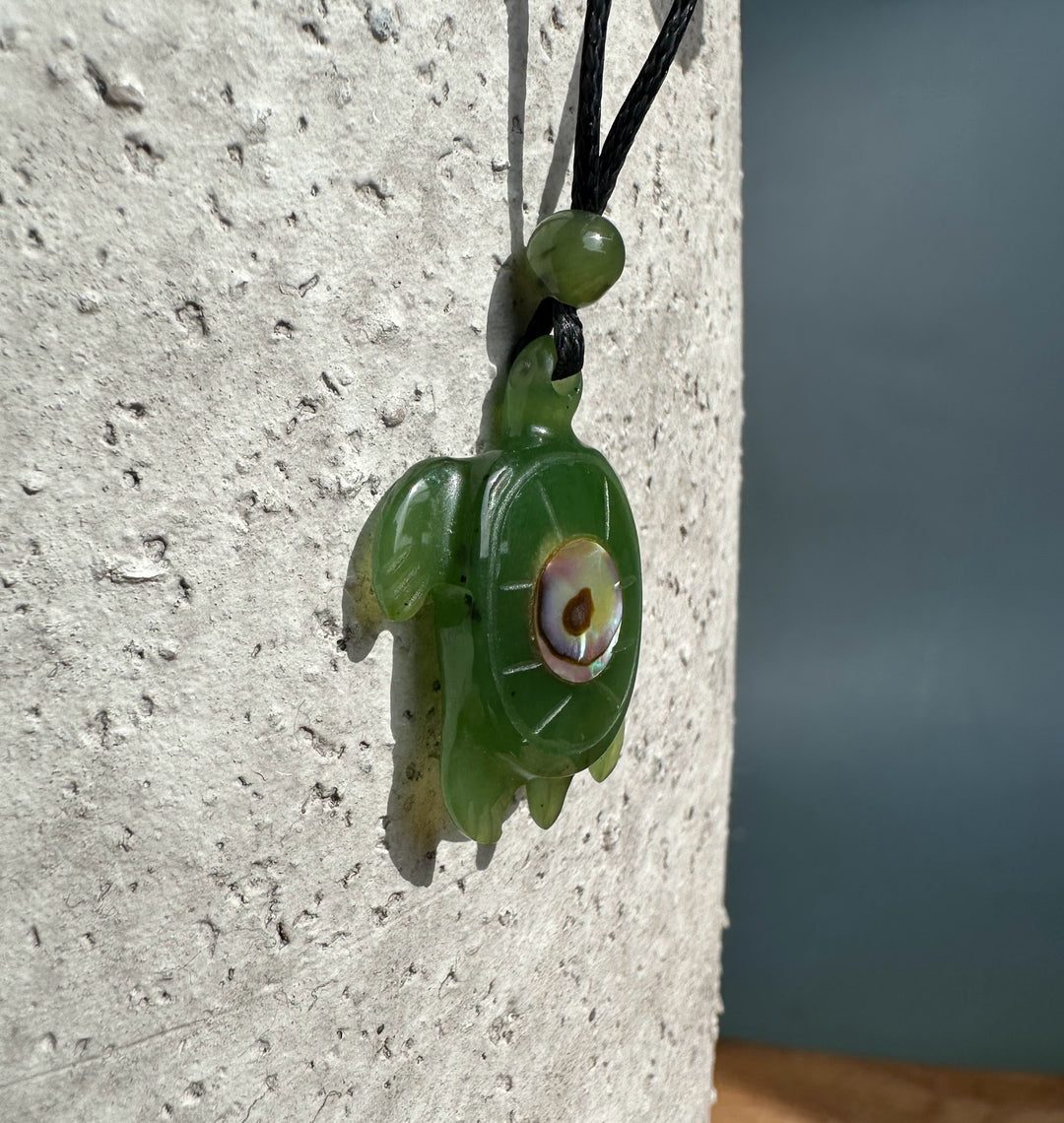 Close up of jade turtle pendant with abalone eye on a black cord against a pale textured surface