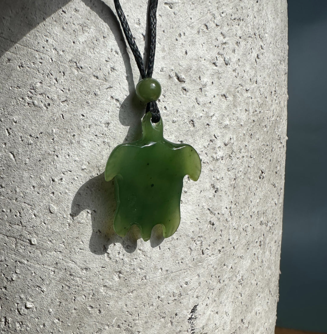 Green jade turtle pendant with abalone eye hanging on a black cord with a bead against a light textured wall