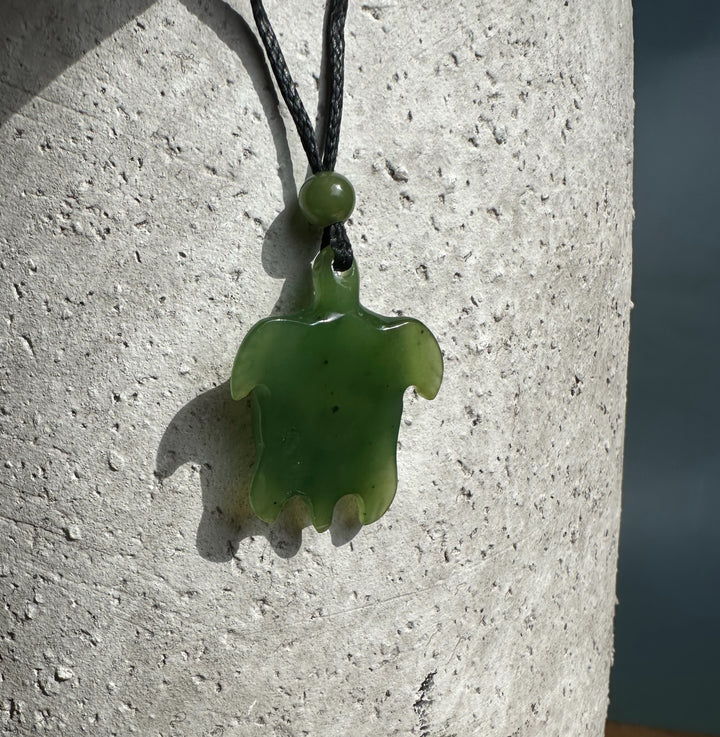 Green jade turtle pendant with abalone eye hanging on a black cord with a bead against a light textured wall