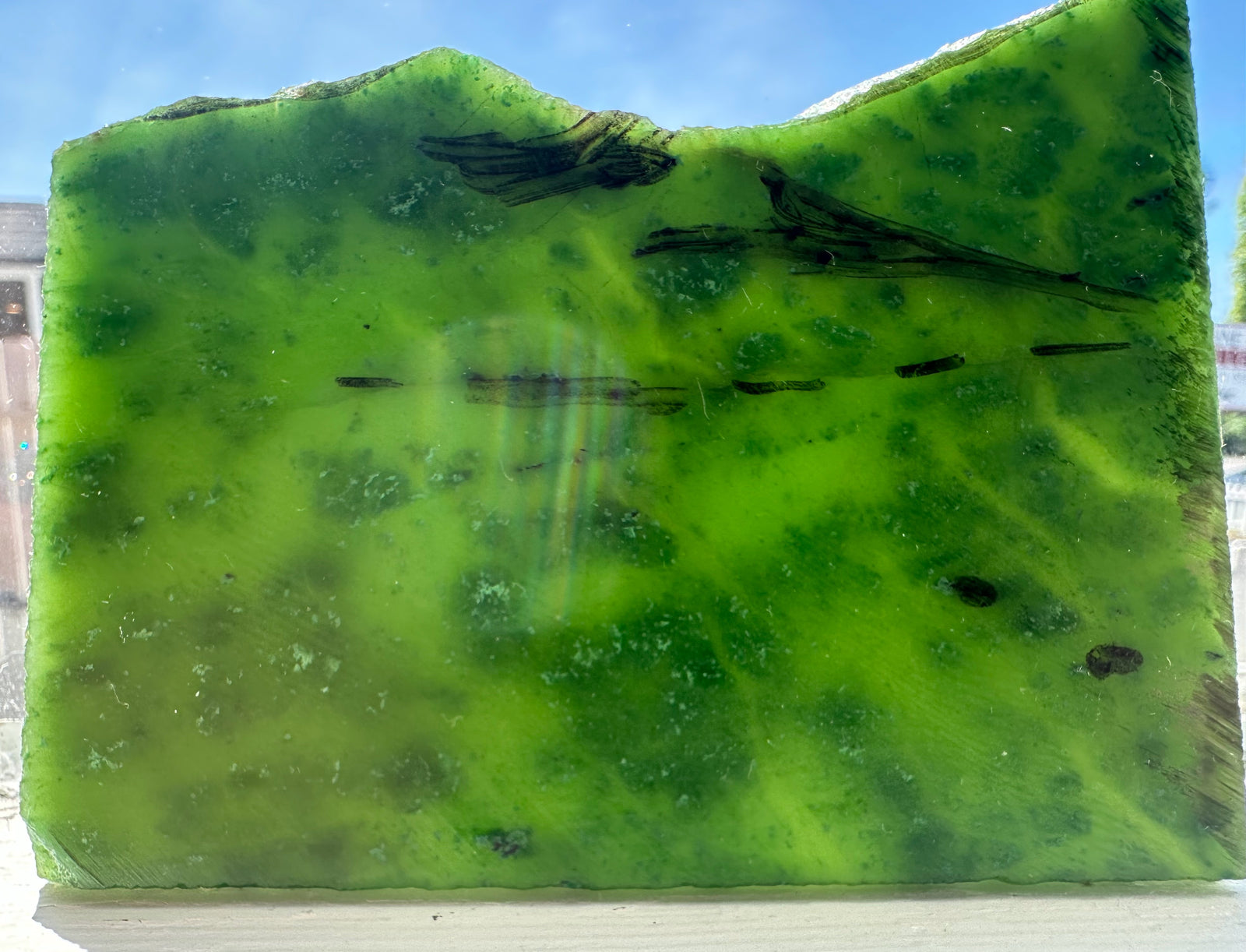 Bright green Cassiar Jade slab with dark inclusions shown outdoors against a blue sky.