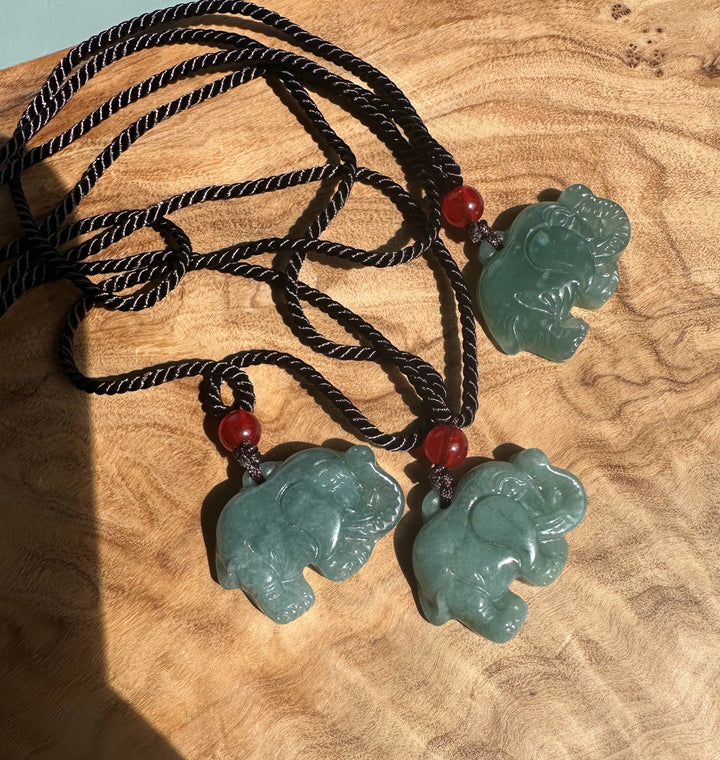 Three jadeite elephant pendants on braided black cords with red beads on a wooden surface