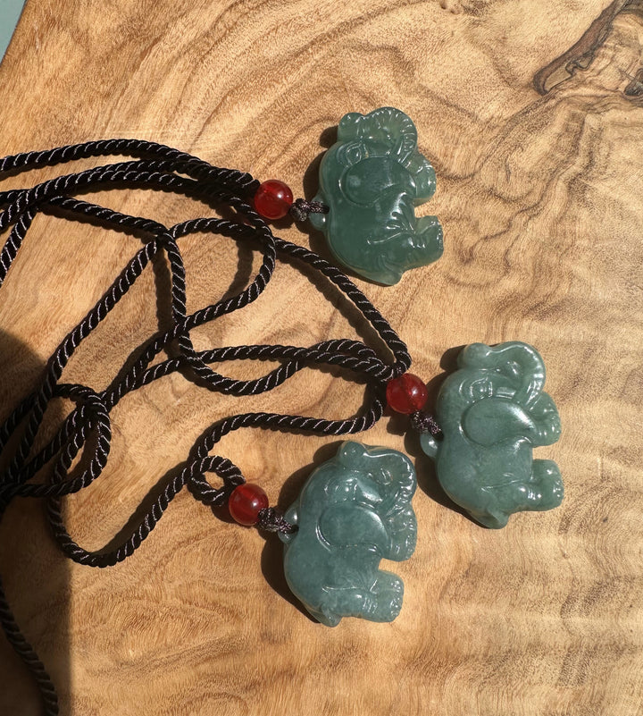 Three jadeite elephant pendants on braided black cords with red beads arranged on wood