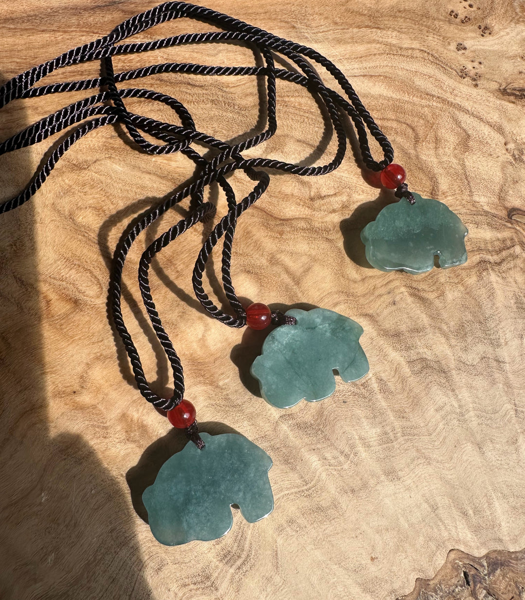 Guatemalan jadeite elephant pendants on black braided cords with red beads laid on a wooden surface