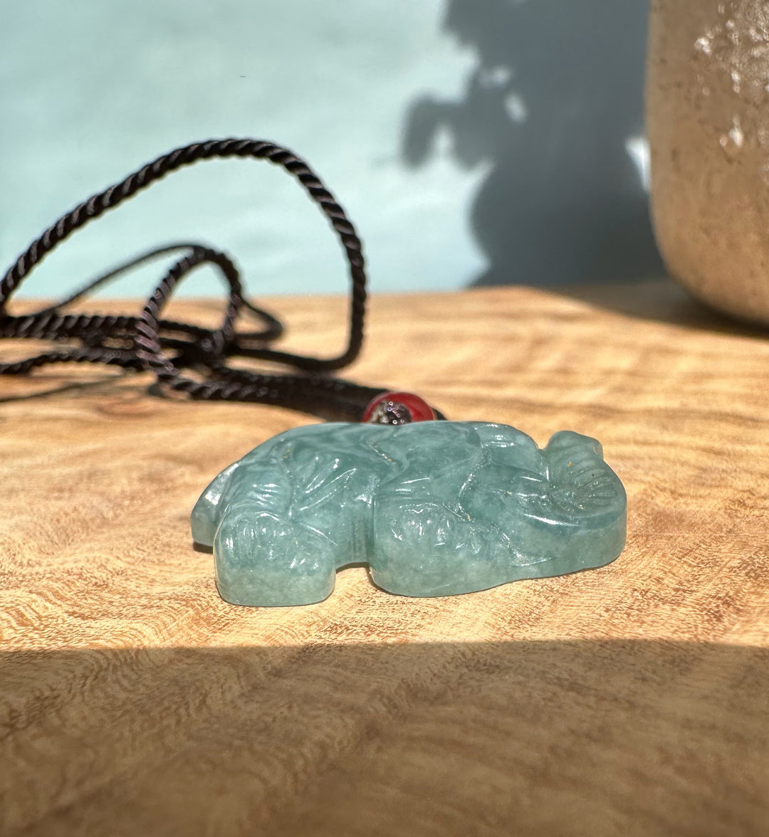Close up of a jadeite elephant pendant on a black cord with a red bead against a wooden background