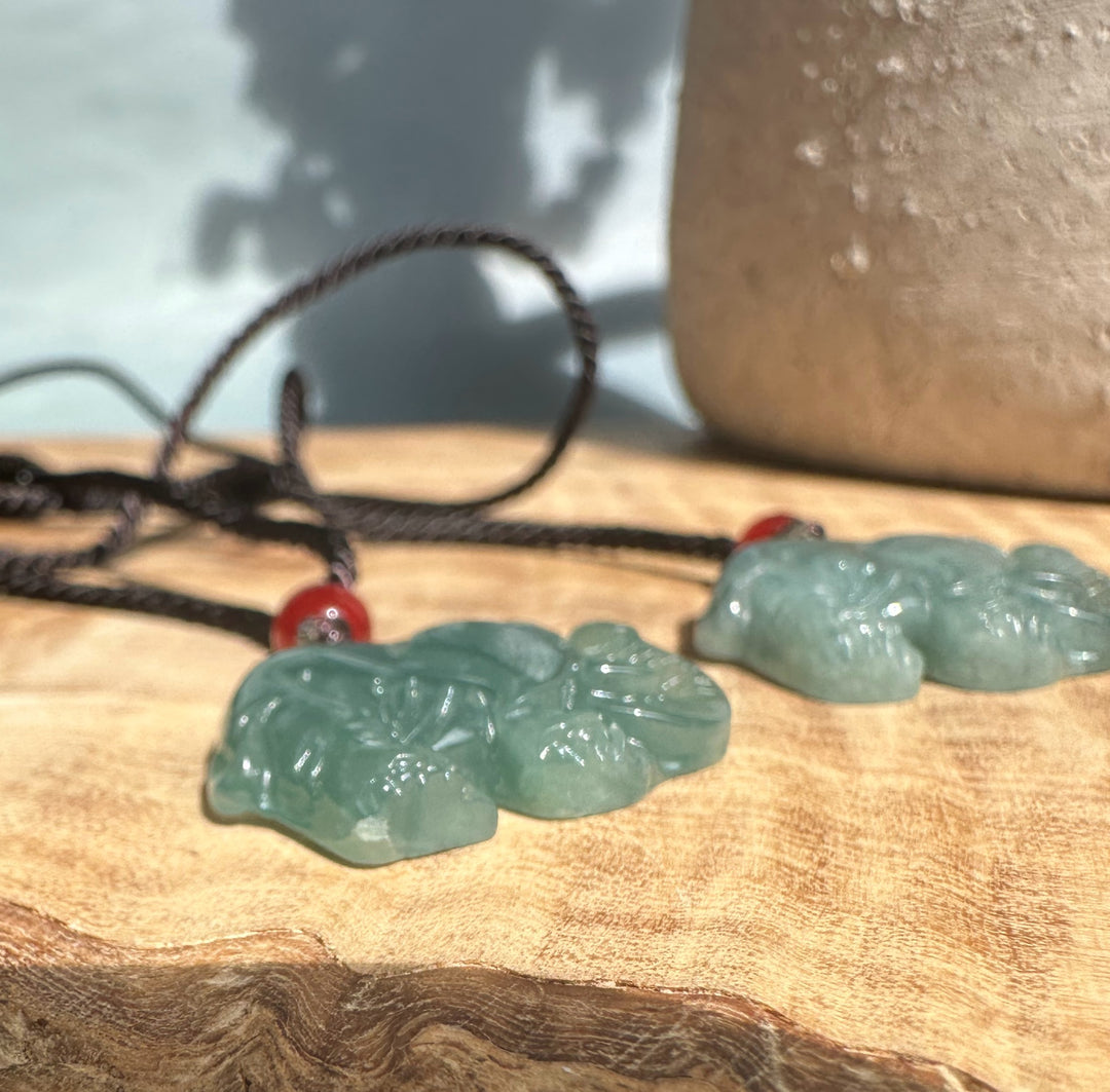 Two jadeite elephant pendants on black cords with red beads resting on a wooden surface