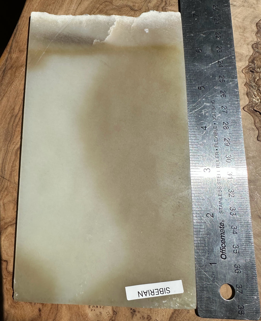 Honey white nephrite jade slab with a rough top edge on a wood surface beside a metal ruler for scale