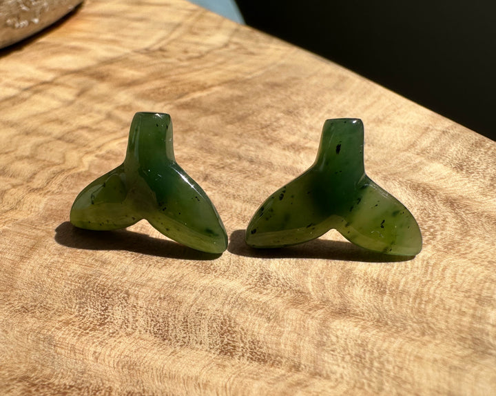 Two green jade whale tail stud earrings resting on a wooden surface.