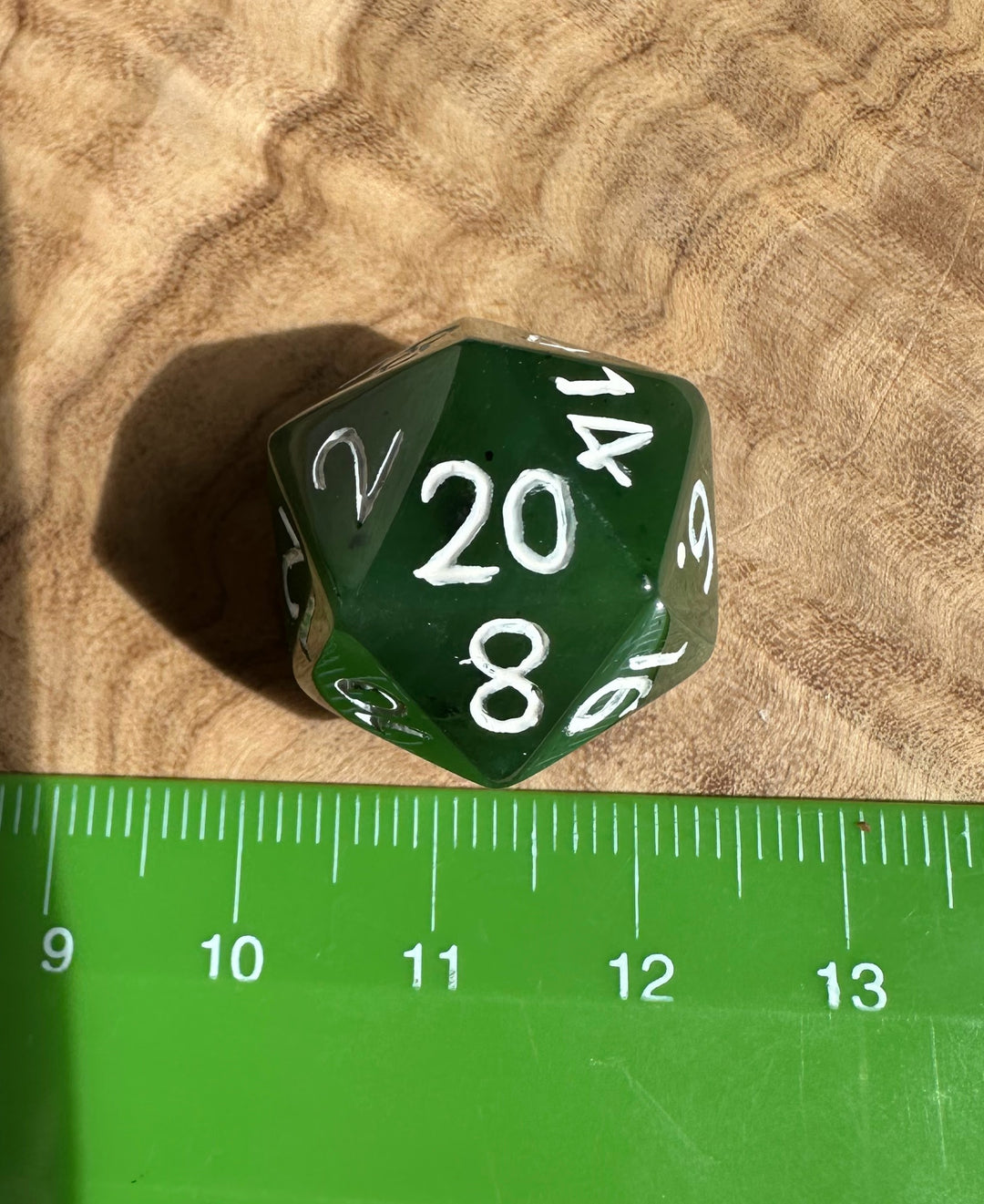 Jade green twenty sided die on a textured wood surface with a green ruler beneath